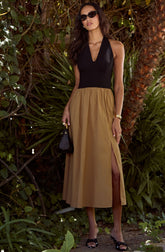 A woman is wearing a sleeveless dress with a black fitted top that has a deep V neckline and a tan, high-waisted, ankle-length skirt with a slit on one side. She accessorizes with dark sunglasses, gold bracelets, and holds a small black handbag. She is also wearing black open-toe high heels.
